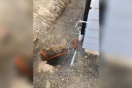 A hole in gravel with white and orange conduit beside a weatherboard wall