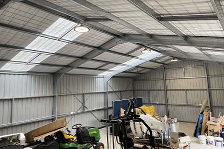 A large steel shed interior with skylights, high-bay lights, a ride-on mower and clutter