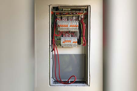 An open flush-mounted switchboard in a wall recess, with breakers and red cables